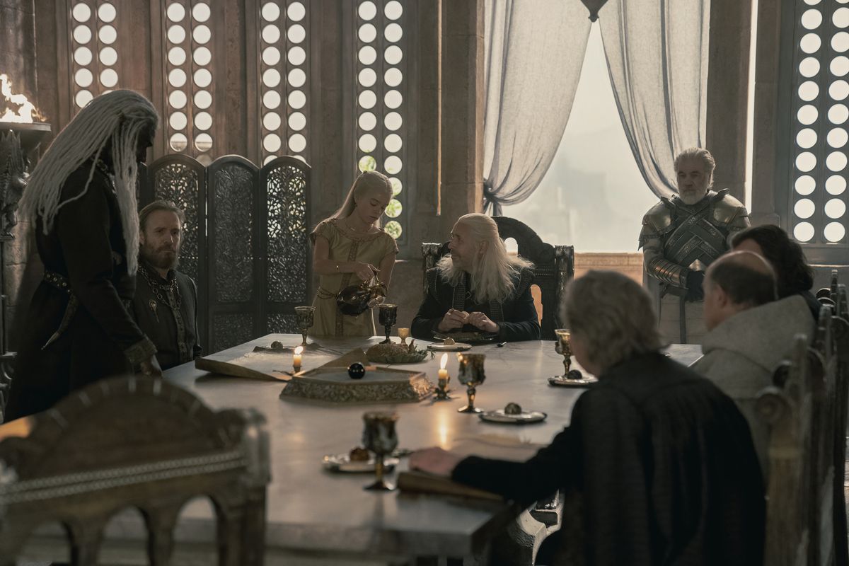 Viserys sitting at his council table with his council around the table and a guard behind him at the window. His daughter is pouring him some wine and he’s looking at her