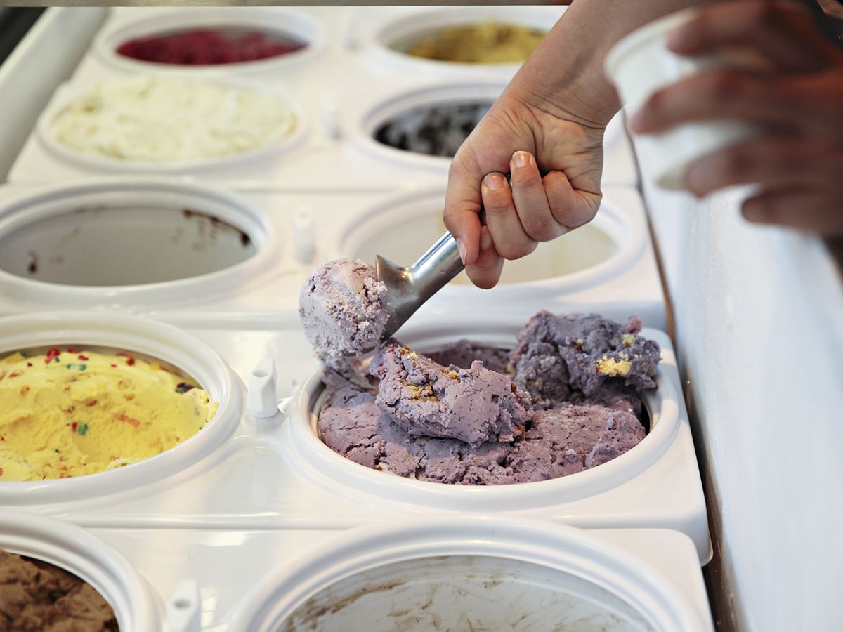 Two lines of ice cream buckets; a hand is scooping a purple berry flavor.