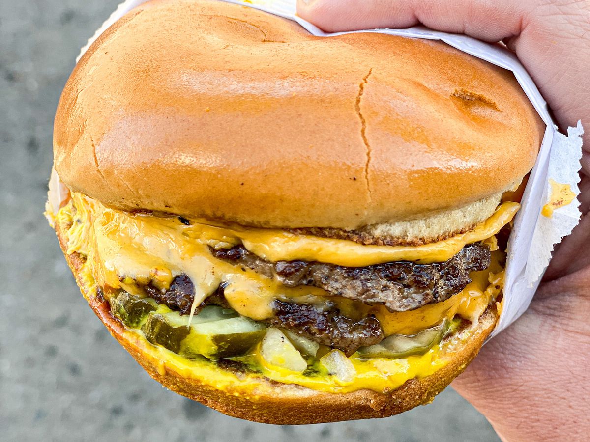 A hand holds a two-pattied cheeseburger with pickles and cheese at daytime.
