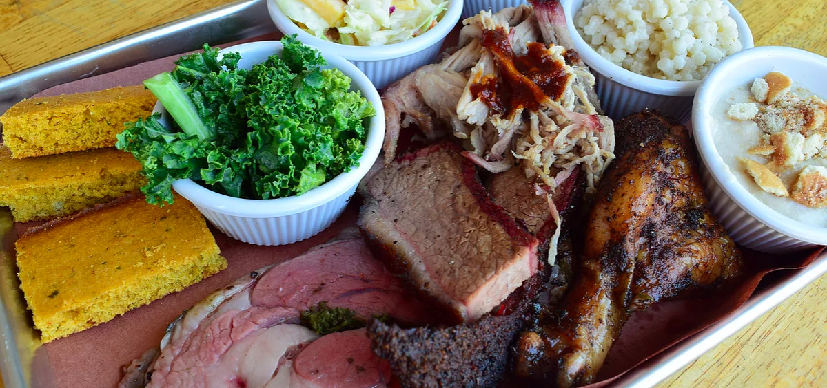 A metal tray on a wooden surface is packed with barbecue: sliced brisket, a smoked turkey leg, pulled pork, and several sides.