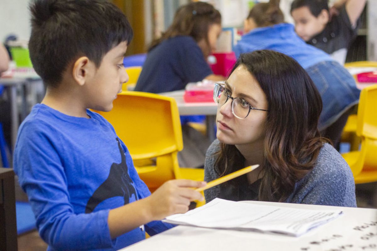 Teacher Sarah Schielke, wearing glasses and blue top, crouches down to listen to a student, who is sitting at a desk and holding a yellow pencil.