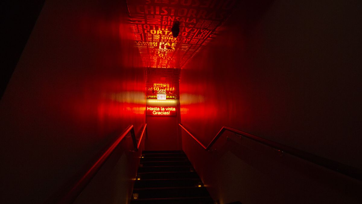A dark stairwell leads up to a glowing, red-lit door with a red neon sign that reads “Hasta la vista. Gracias!”