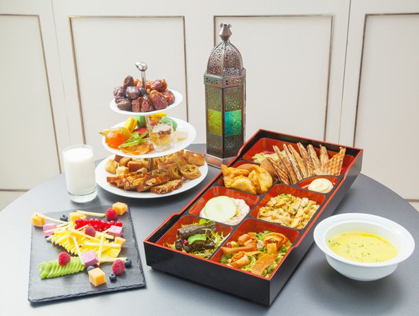 A black box with portioned containers of meets, veggies, salads, and desserts, plus fruit and cheese on a small tray, a bowl of soup, and a three-tiered tray with Mediterranean desserts.