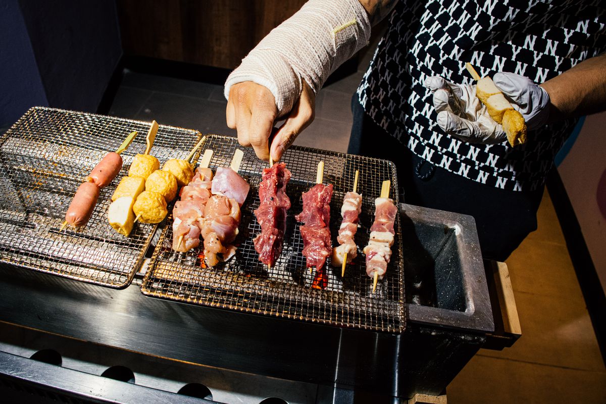 A person wearing a cast tends to a charcoal grill station laden with skewers.