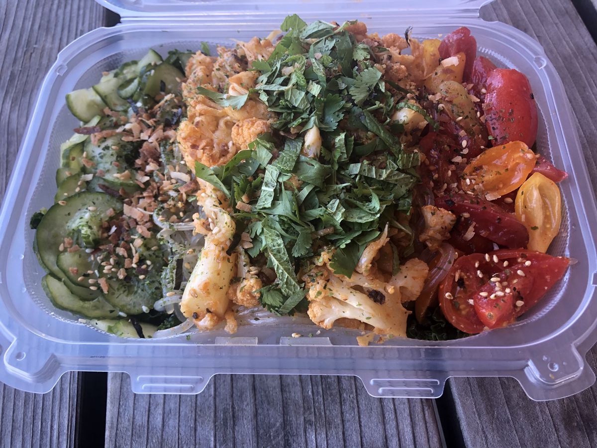 A takeout container is filled with red-hued cauliflower, sliced grape tomatoes, and cucumbers covered in furikake at Meals 4 Heels.