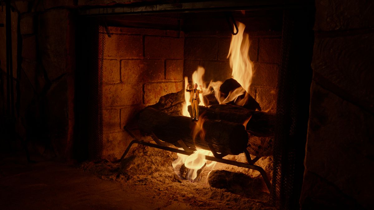 A tiny man in a fancy suit stands on top of the front log of a roaring fire in Adult Swim’s yule log horror movie The Fireplace