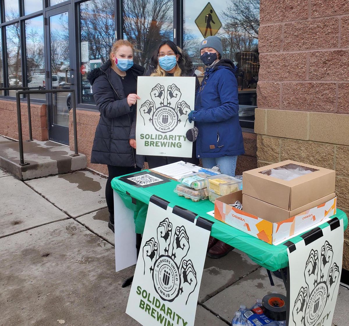 Three young people wearing coats and facemasks stand outside a Starbucks building holding a “solidarity brewing” poster with four raised fists on it.