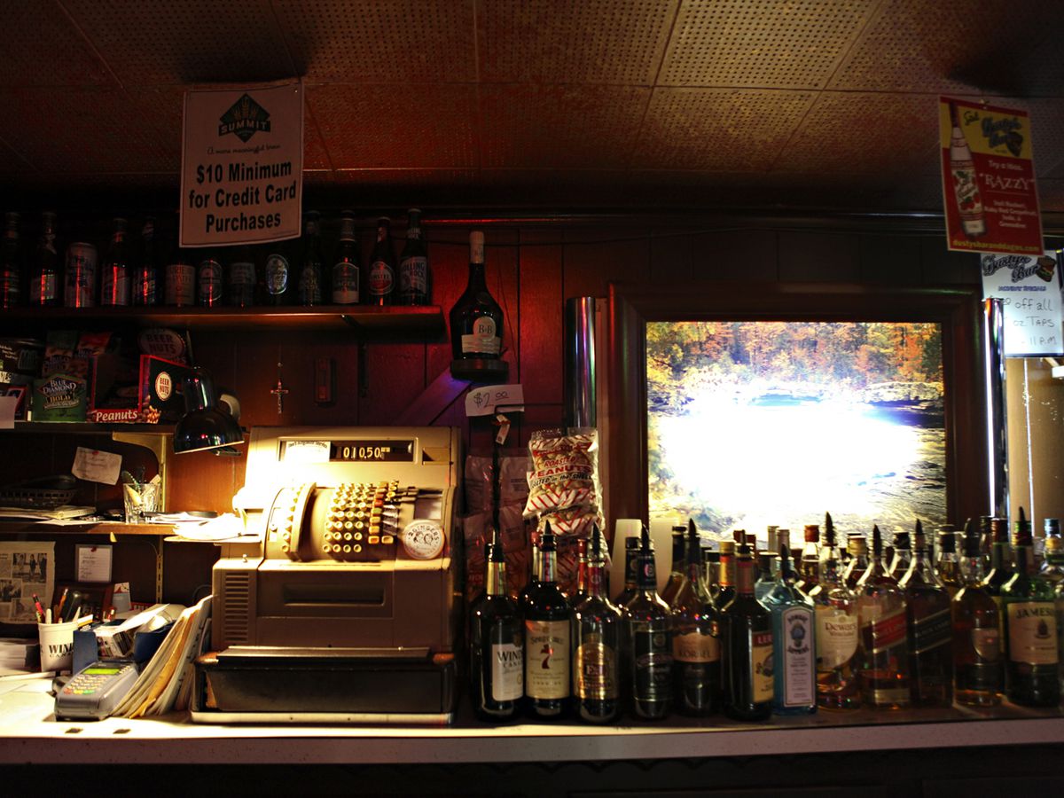 A counter behind a bar set with many liquor bottles stacked next to one another, and an old-school cash register next to it.