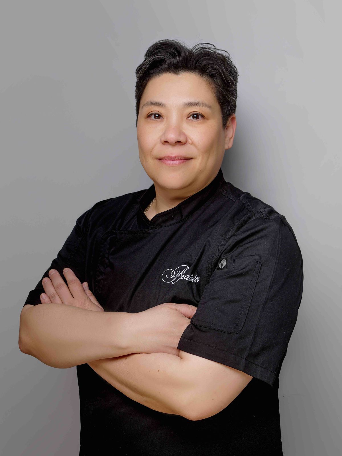 A woman stands in a short sleeved, black chef’s top, with Beatrice embroidered on it in white.