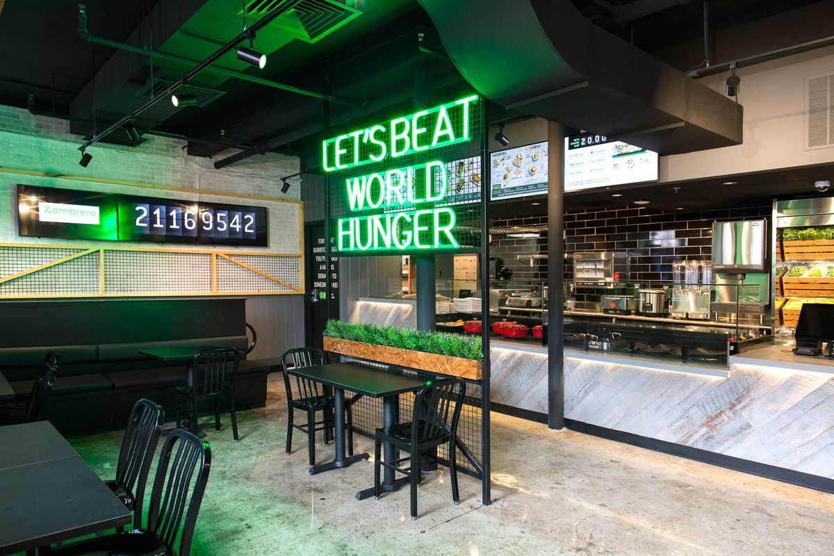 A neon sign reading “let’s end world hunger” hangs in front of an ordering counter inside Zambrero