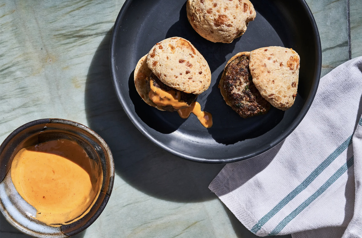 Lamb sliders sit inside a Brazilian cheese bread sitting in a black ceramic bowl above a marble counter top and a cloth napkin. 