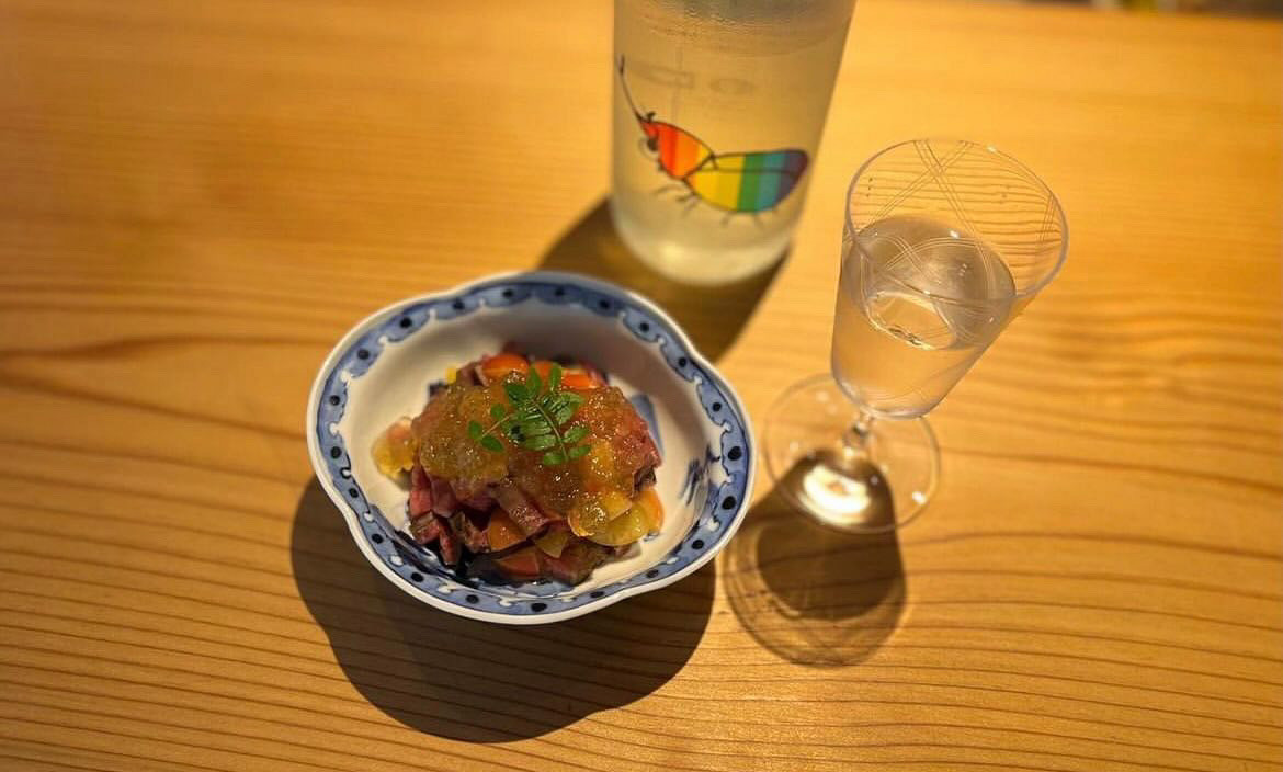 A small snack in a decorative dish, beside an ornate glass of sake and a bottle.
