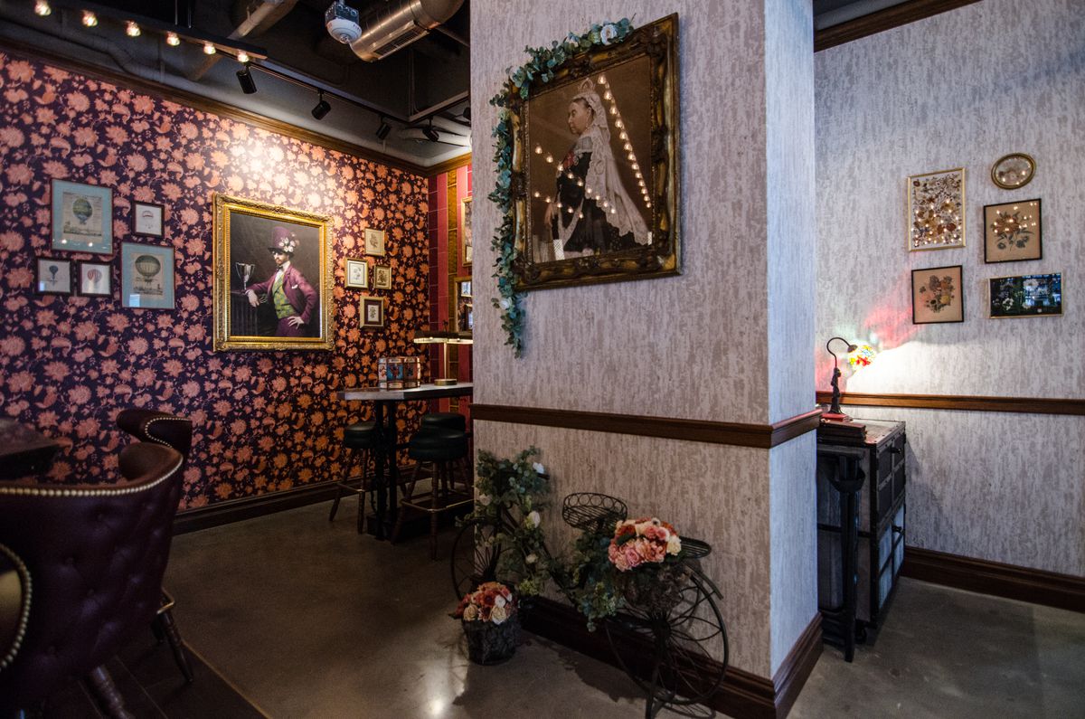 Interior of a restaurant featuring a variety of Victorian-inspired artwork. One wall has dark floral wallpaper. There’s a decorative black wire bicycle covered with flowers and ivy parked against a wall.