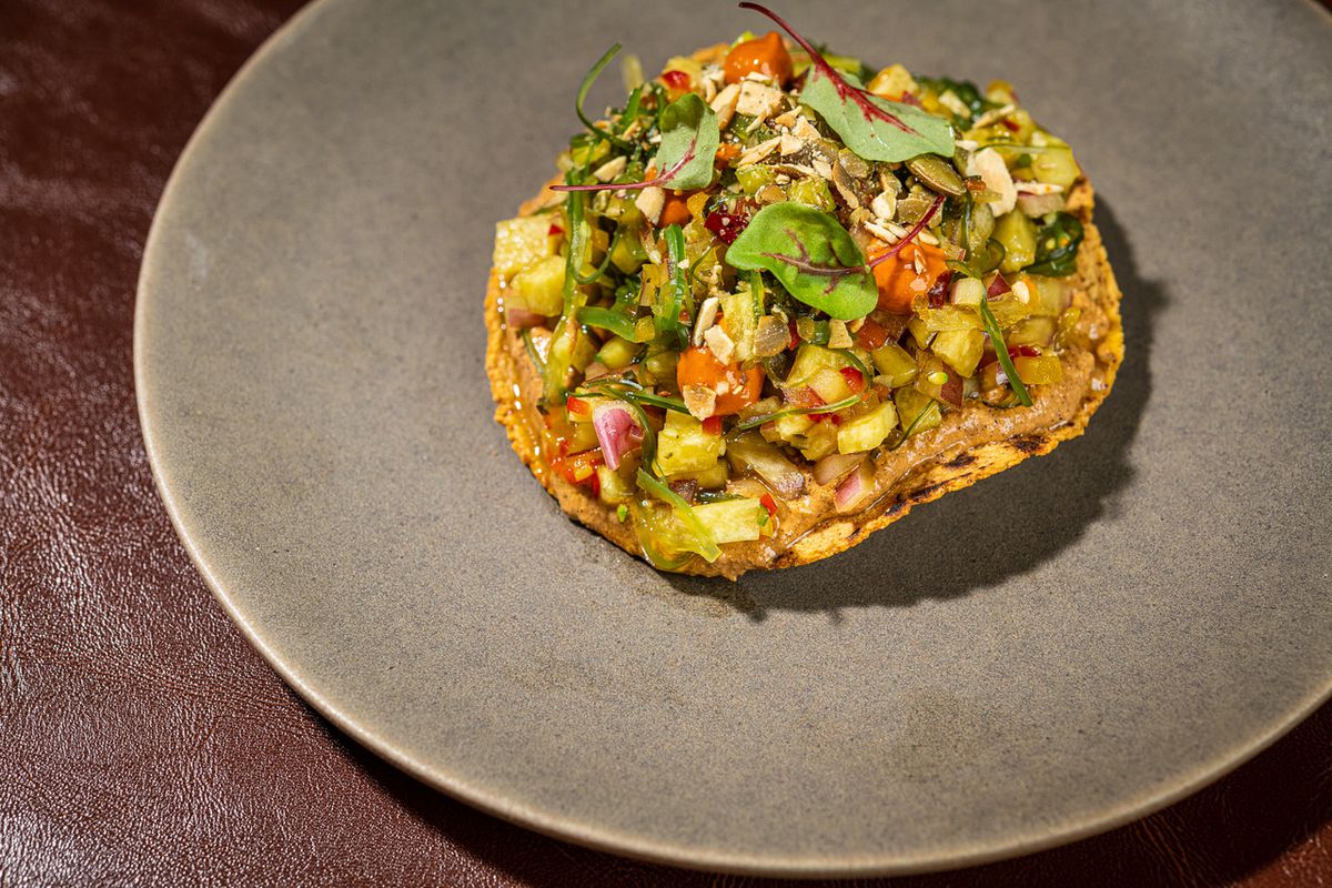A close-up of a yellow corn tostada topped with a Yucatan pumpkin seed sauce and a confetti of crisp produce (jicama, bell peppers, seaweed, cucumber, red onion) with a red chile emulsion, salsa macha, and cilantro