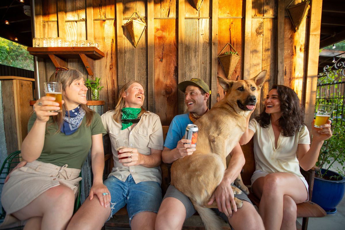 A group of four people and a dog sit on a bench drinking beers.