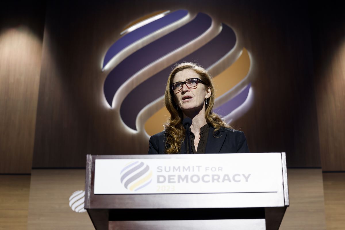 Samantha Power, a white woman with glasses and long brown hair, wearing a dark suit, speaks from a podium with the logo of the Summit for Democracy behind her.