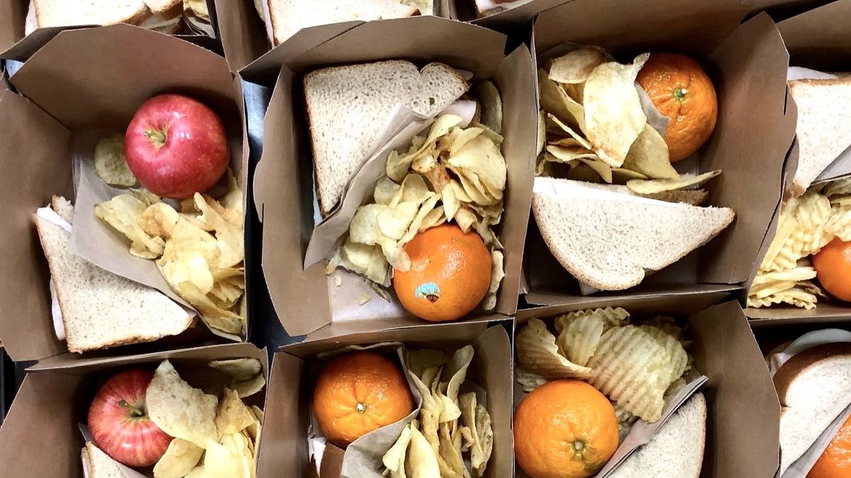 Brown takeout containers filled with crinkle cut chips, oranges, apples, and sandwiches sliced in half.