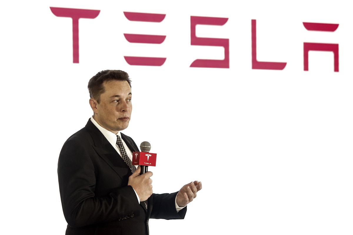 Elon Musk speaks into a handheld microphone, wearing a dark suit and tie and standing in front of a white backdrop with a large red Tesla logo.  