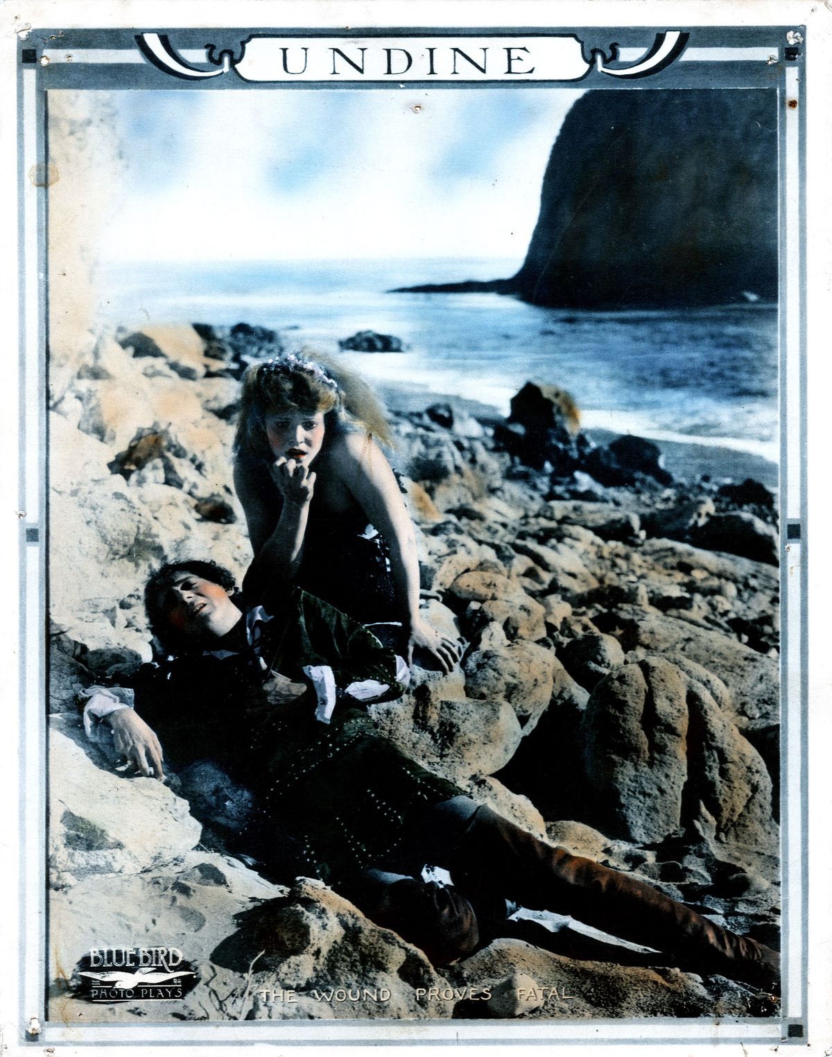 An antique colorized photo shows a young woman crouched over the body of a man, among rocks at the edge of the sea.