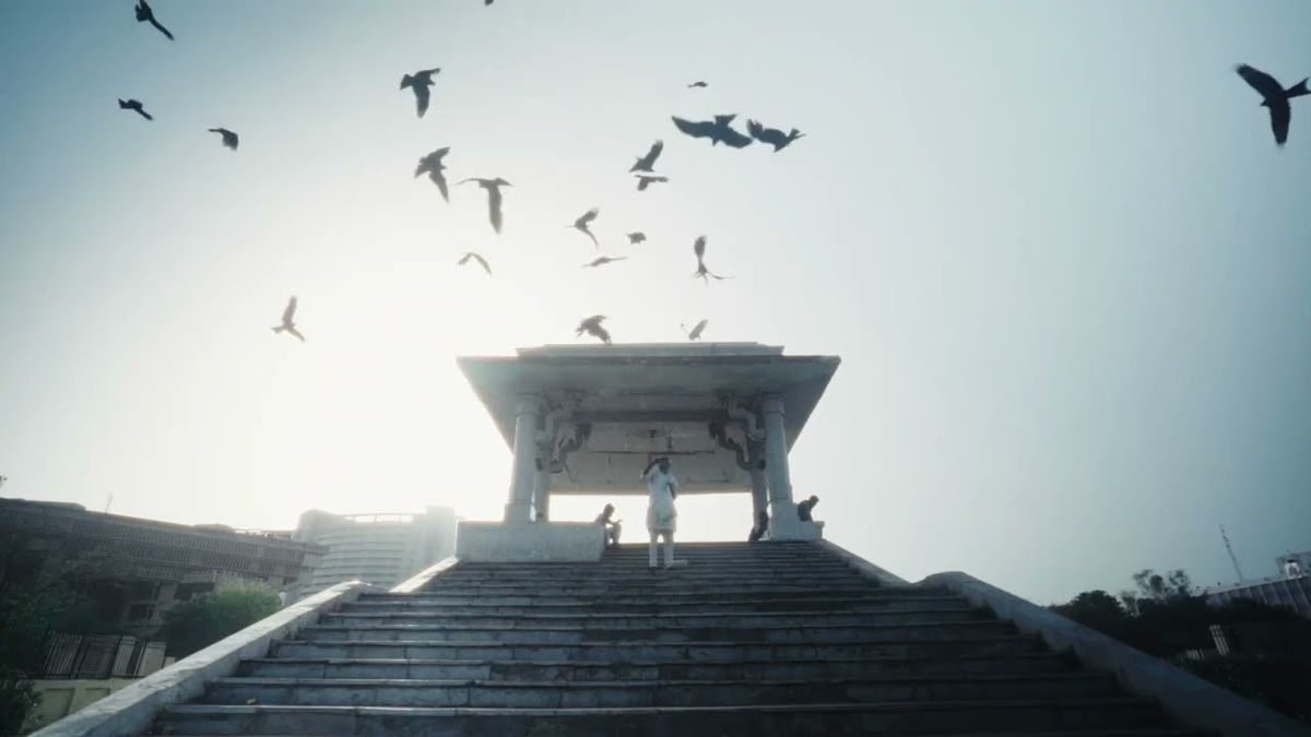 Birds fly overhead while a person in white looks up at them on a set of stairs, leading up to a white marble column, in All That Breathes.