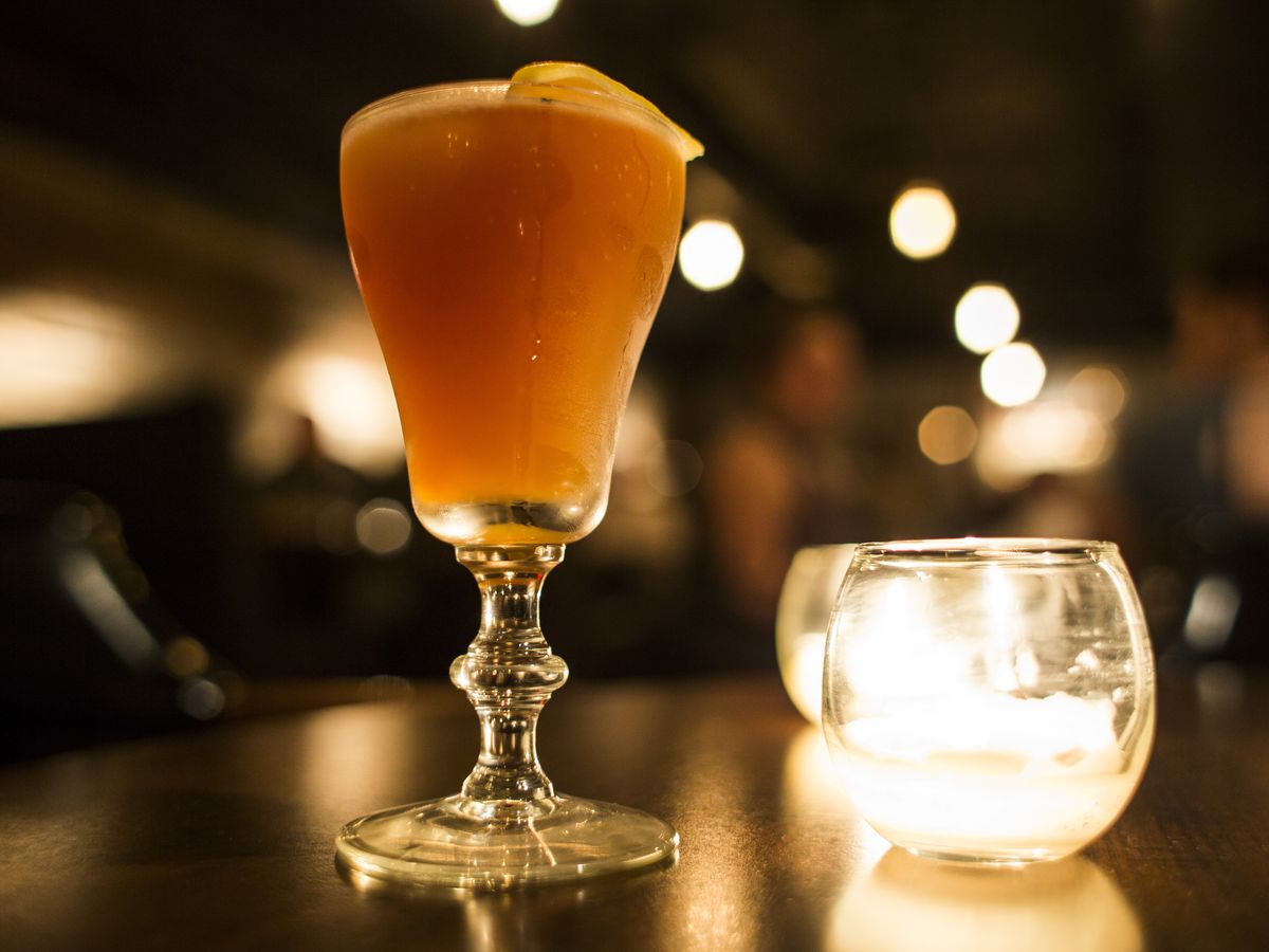 The orange colored cocktail sits next to a glass candle holder on a wooden table in a dimly lit bar.