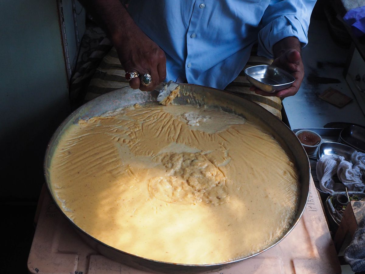 A vendor scoops a thin layer from a massive pan of kheer into a small bowl.