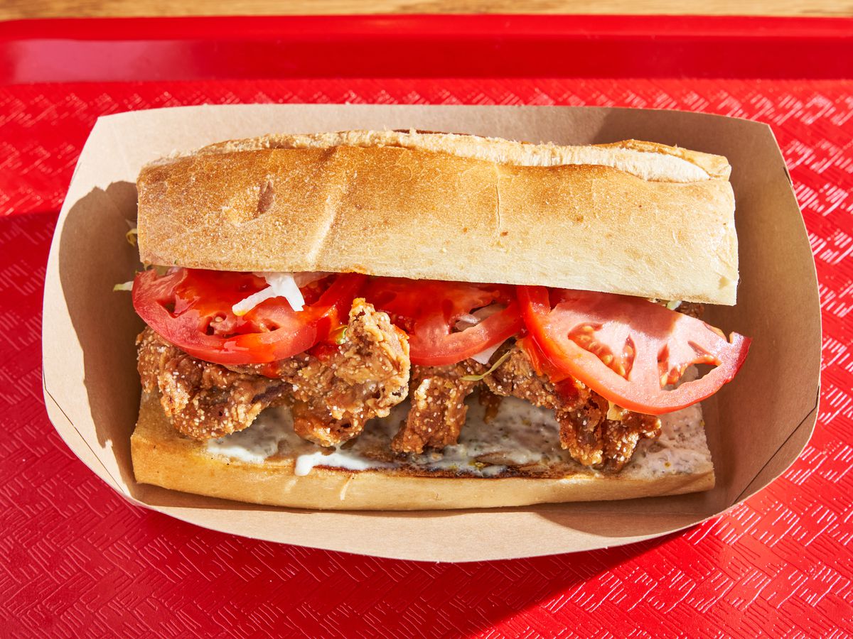 A sandwich with fried oyster mushrooms and tomato is strewn out on a sliced hero roll in a paper boat on a red cafeteria tray.