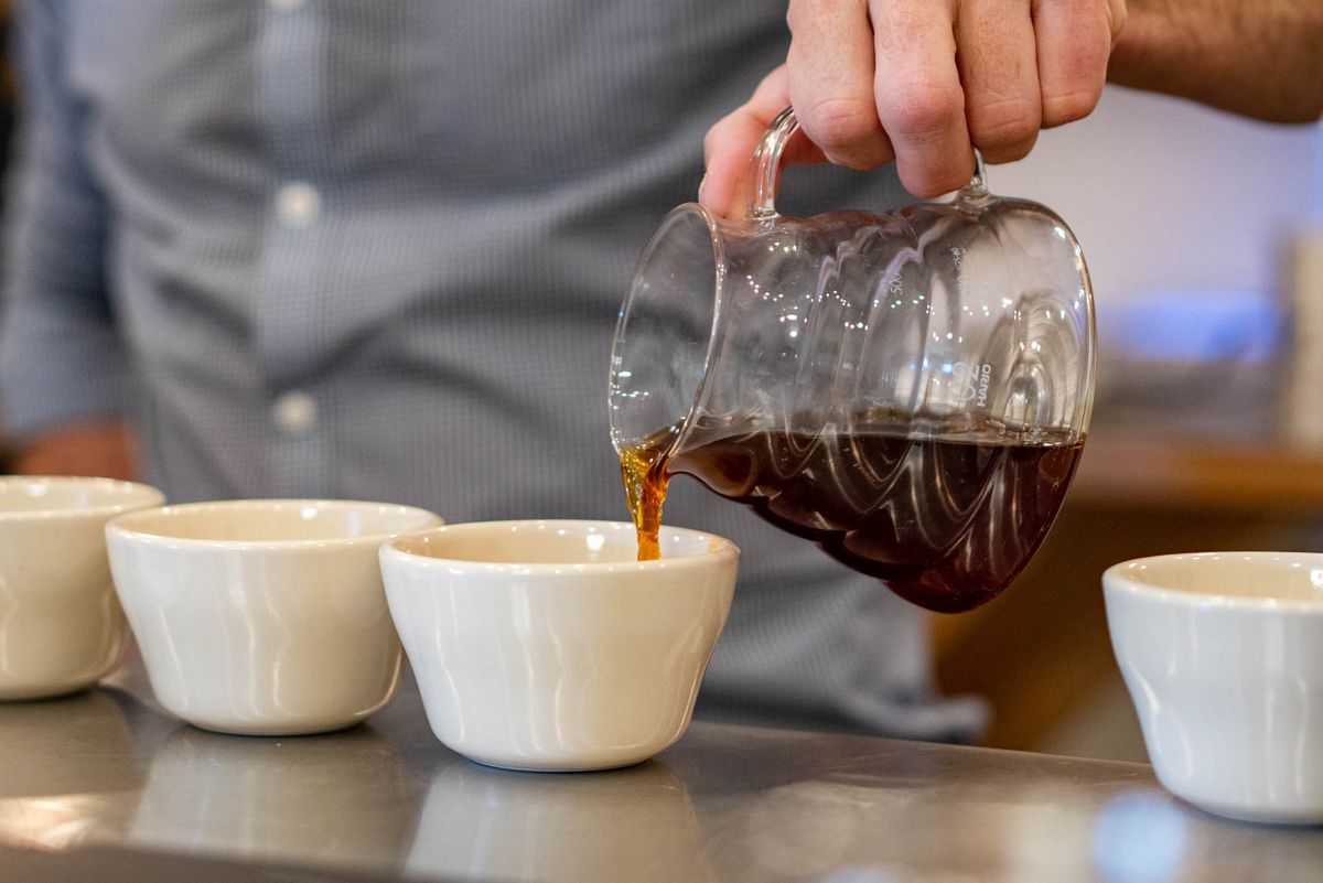 Blas Alfaro pours coffee from a glass pitcher into one of four ceramic cups lined up on a metal countertop.