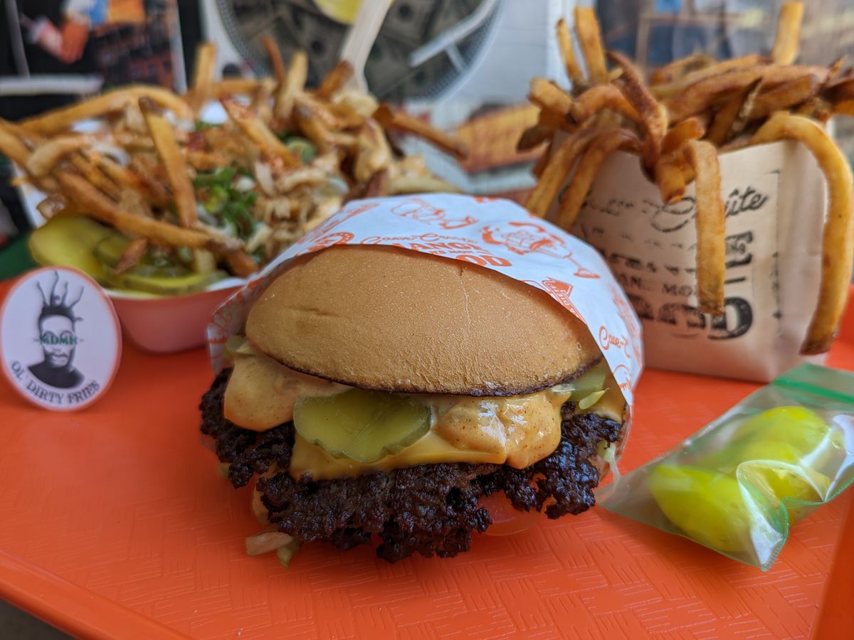 A smash burger with cheese and pickles, on a tray with two bags of fries with various toppings.