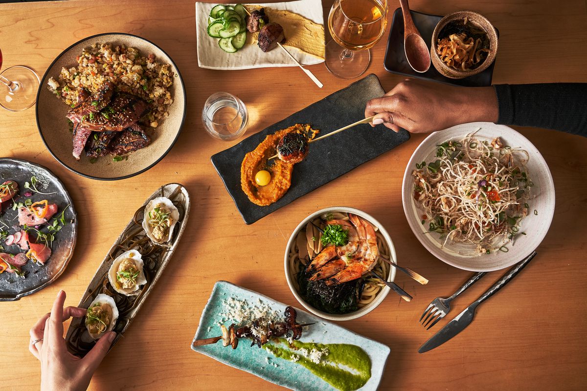 An aerial view of a spread of small plates topped with Asian-inspired fare over a wooden table