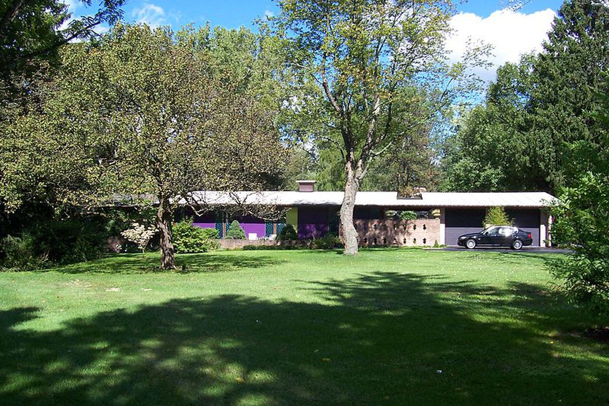 A single-story house clad in colorful aluminum panels and brick sits behind a lush green lawn, tucked into a stand of trees. A car sits in the driveway, in front of the home.