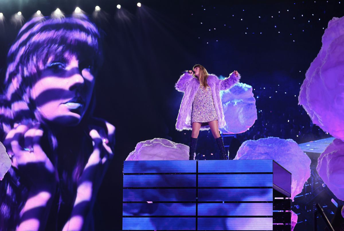 A performer singing wearing a sparkly dress and purple fuzzy coat on a raised platform.
