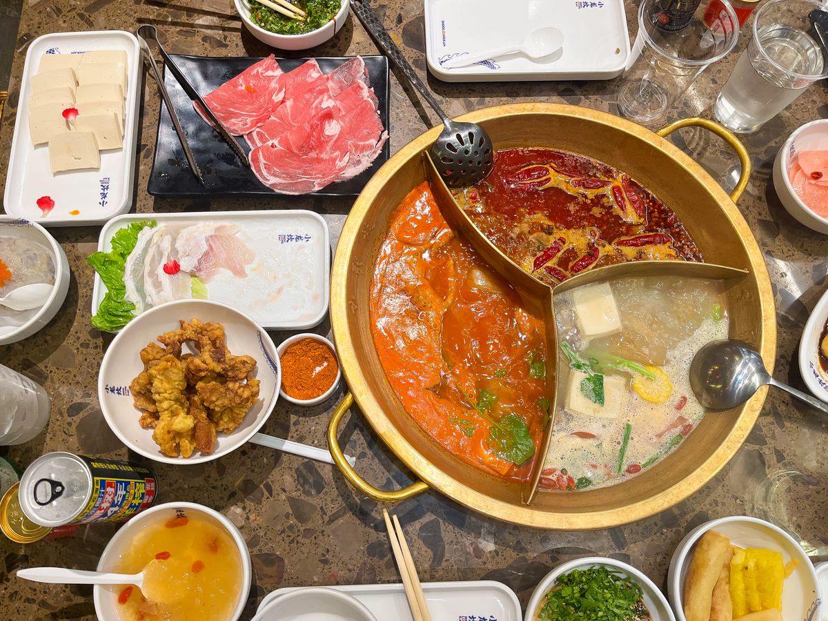 A three-flavor hot pot is surrounded by assorted fixings including tofu and beef slices for dipping.