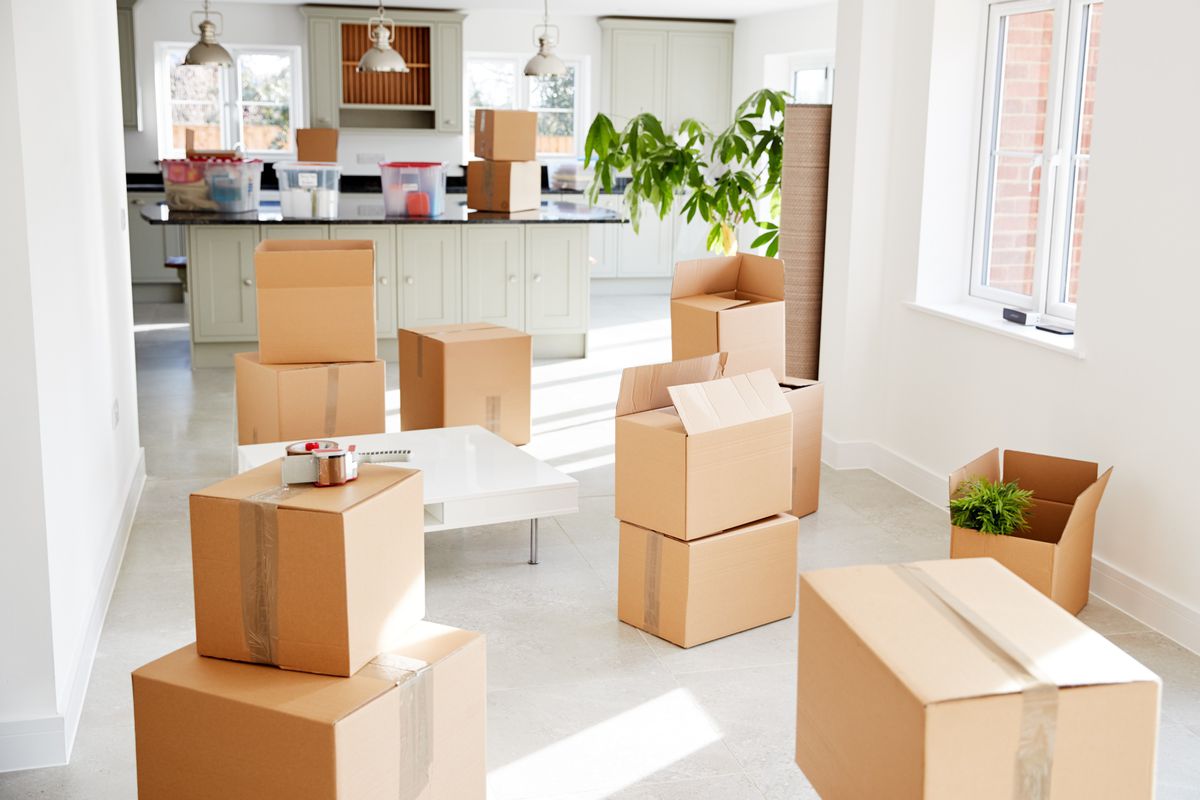 Moving boxes near a kitchen of a home with white walls, white floor, and bright windows. 