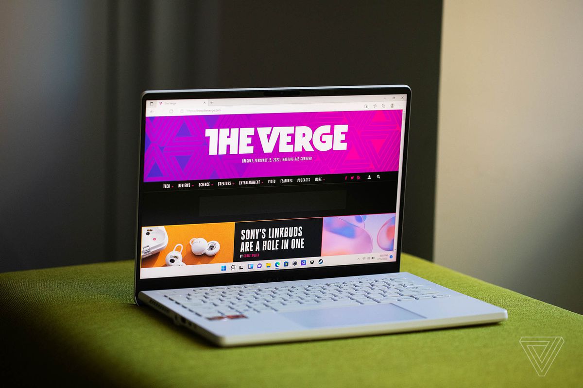 The Asus ROG Zephyrus G14 on a green plush ottoman. The screen displays The Verge homepage.