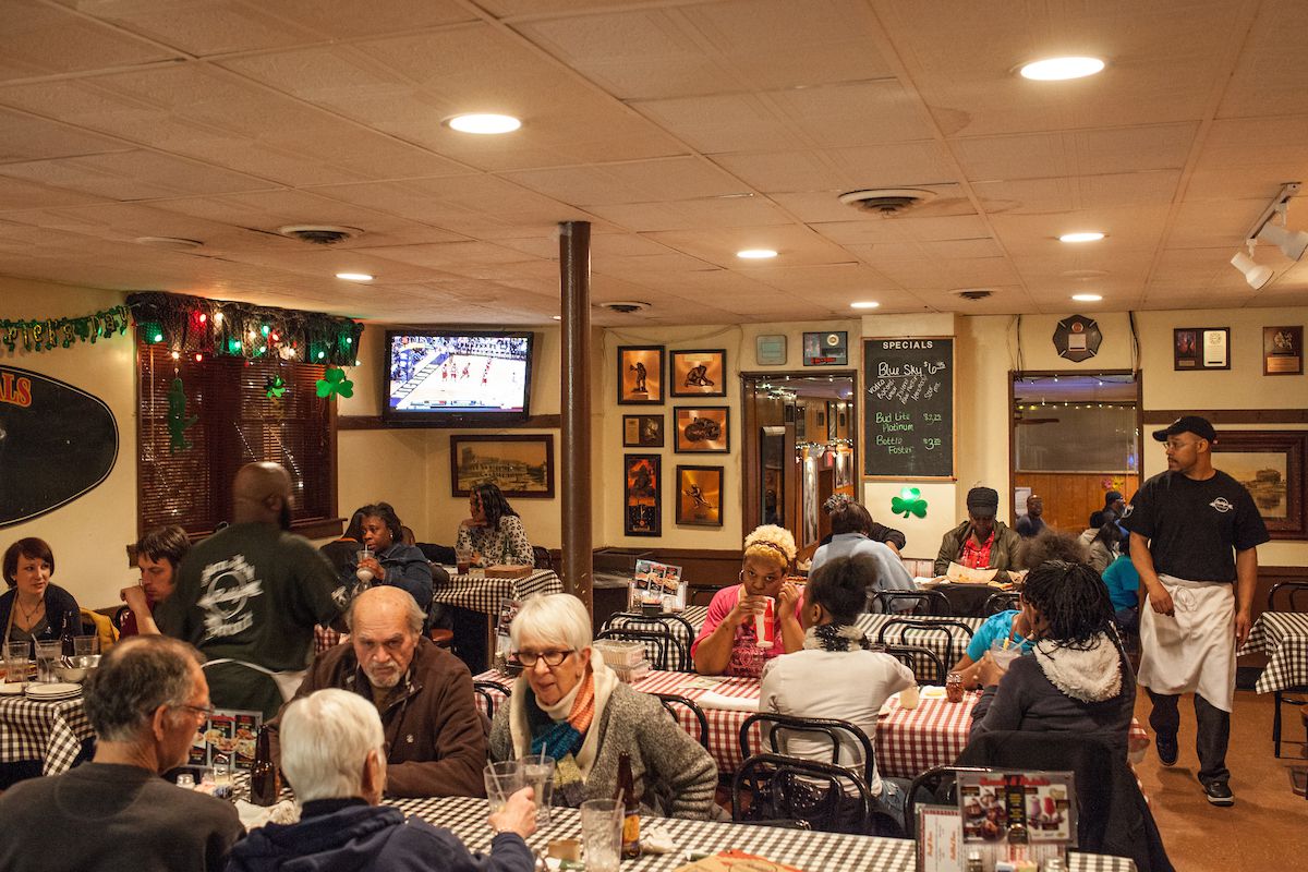 Older customers sit at check tablecloth covered tables below a drop ceiling inside Buddy’s Pizza.