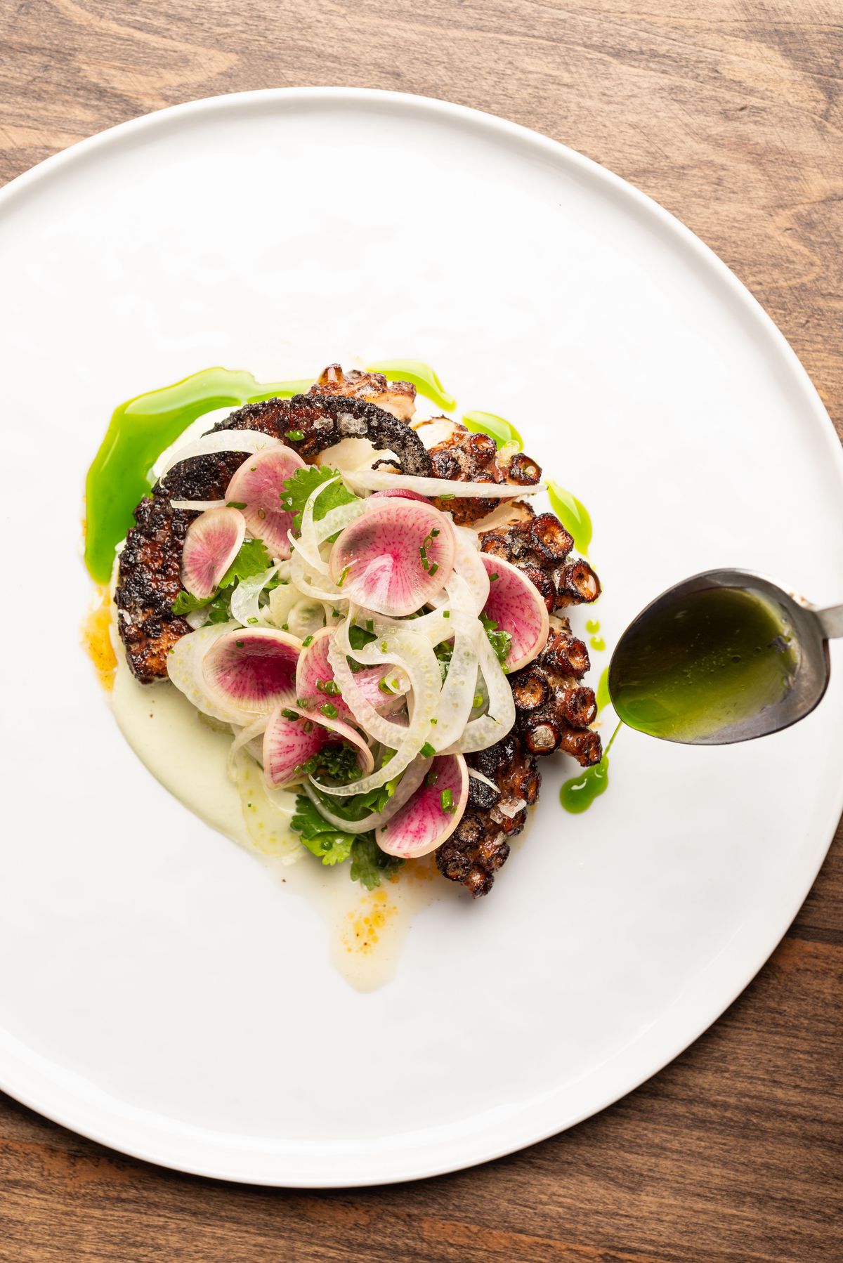 A spoon drizzles olive oil atop an arm of octopus with radishes on top.