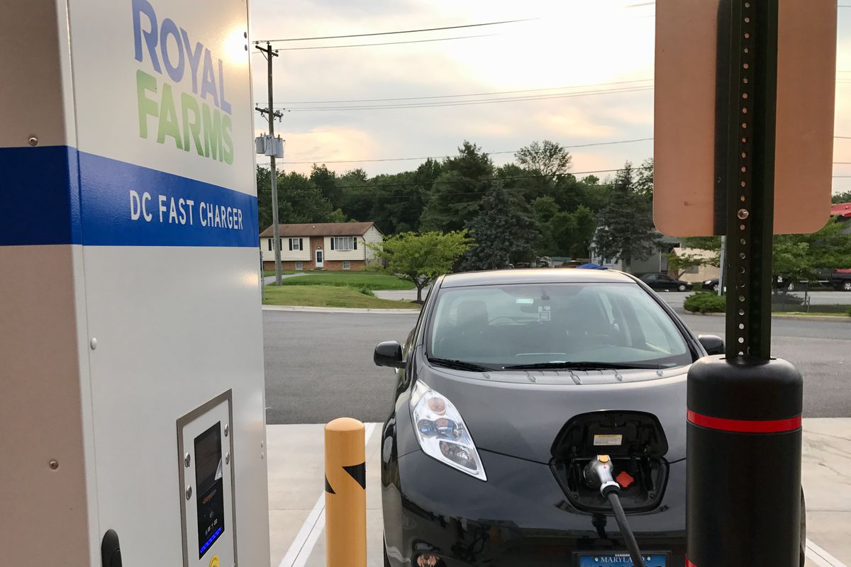 At sunset, a black Nissan Leaf stands headlong towards a Royal Farms DC fast charger.  The CHAdeMO plug is already in the Leaf and its front latch is open.  In the background is a small suburban house with several trees surrounding it, with an empty convenience store parking lot in the foreground.