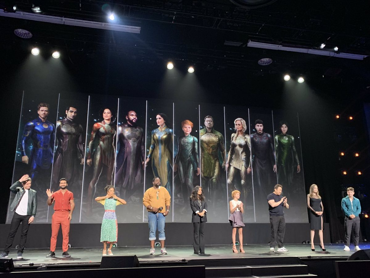 The cast of The Eternals on stage in front of a post of their costumed characters