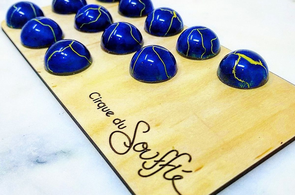 A dozen half-sphere shaped blue bonbons with yellow accents sit on a wooden board branded “Cirque du Soufflé.”