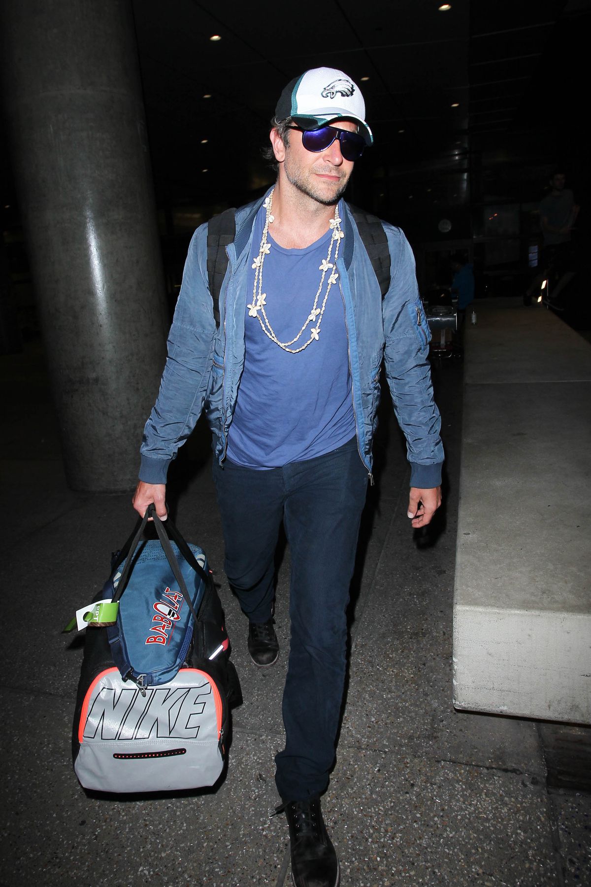 Bradley Cooper walking with a duffle bag and wearing black sunglasses and a white Eagles baseball cap