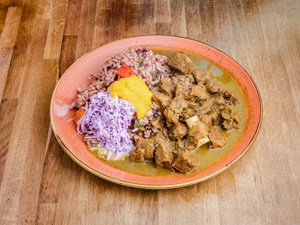 A red plate, piled high with a brown stew, along with Caribbean rice and peas and a finely cut slaw, on a wooden background.