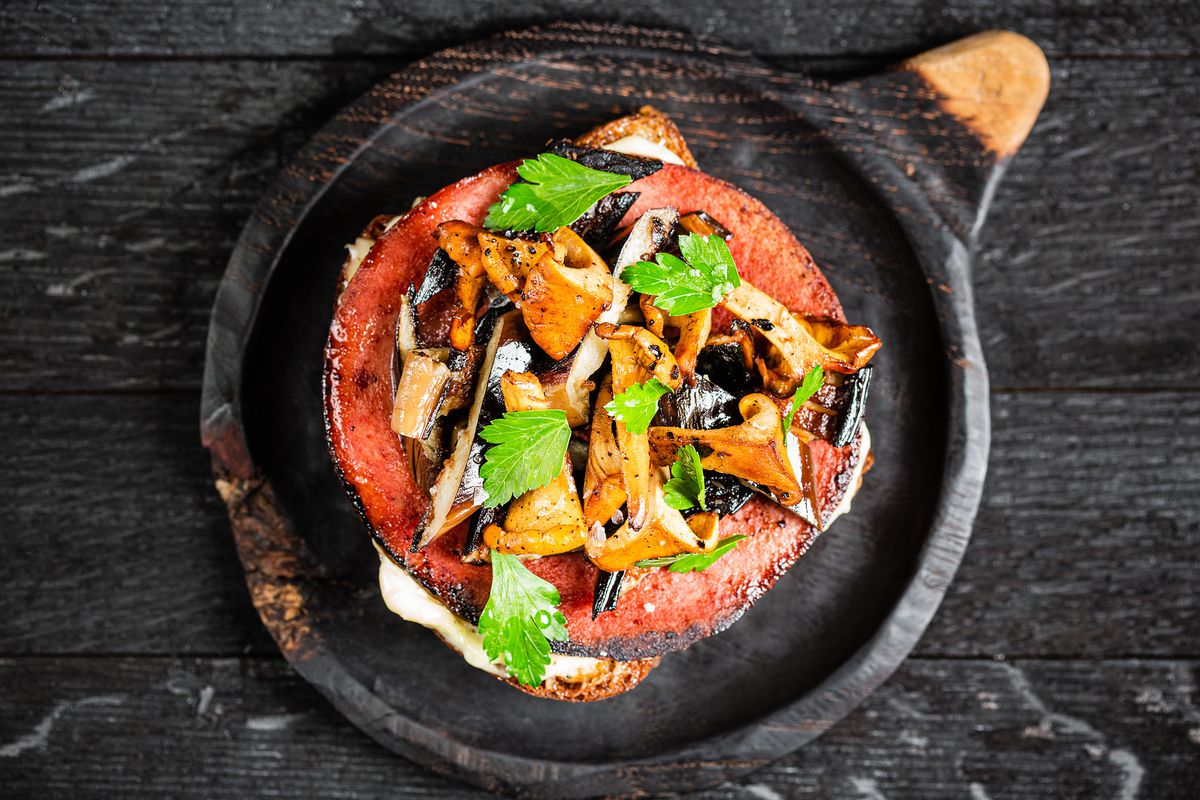 A serving of eggplant, bologna, and mushroom burnt toast, served on a rustic wooden plate.