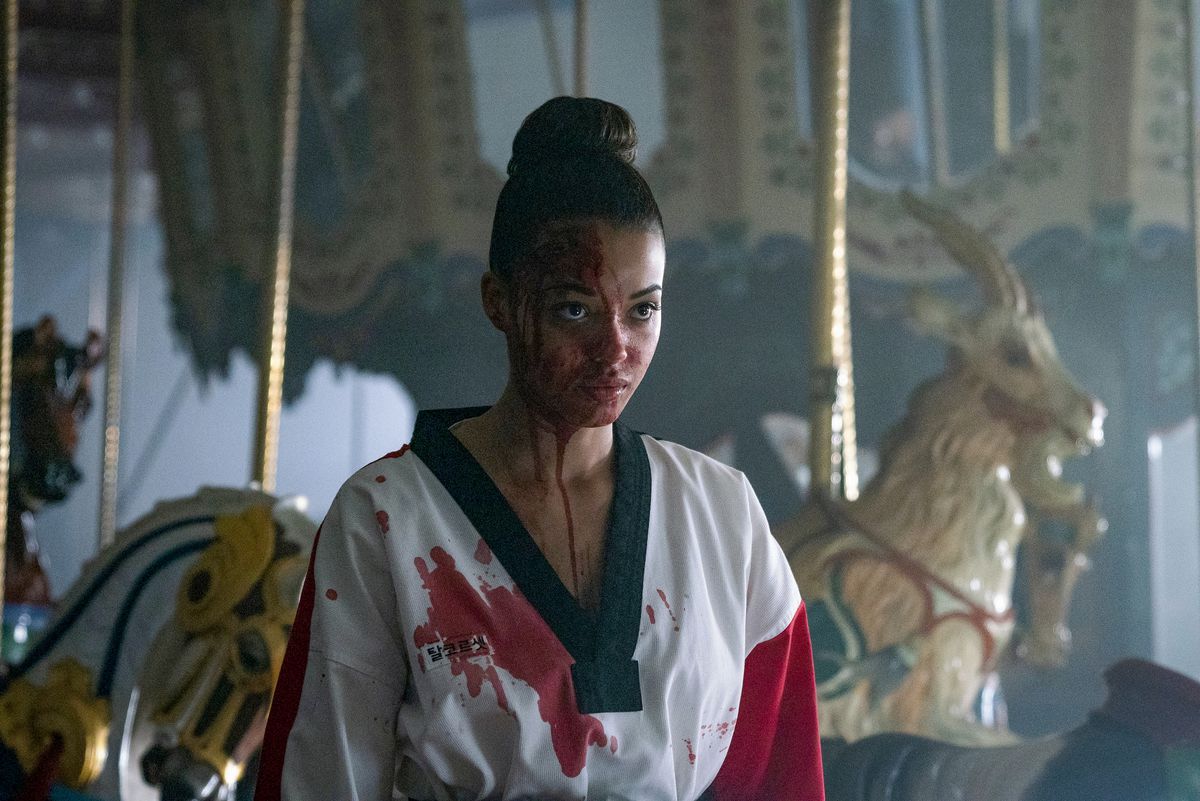 A woman in a bloodied karate uniform stands in front of a carousel, staring blankly at someone out-of-frame.