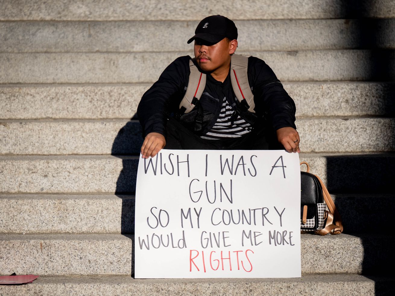 A man in a baseball cap sits on a flight of stairs with a sign propped on his knees reading “Wish I was a gun so my country would give me more rights.”