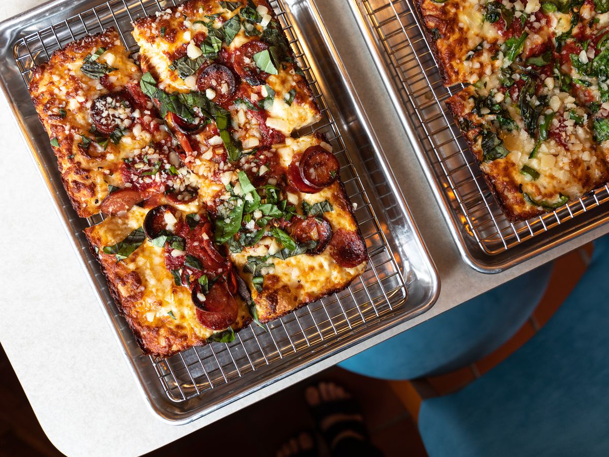 A rectangular pizza cut into four quarters with lots of fresh basil, crumbled cheese, and thick-sliced pepperoni on a cooling rack inside a metal tray. 