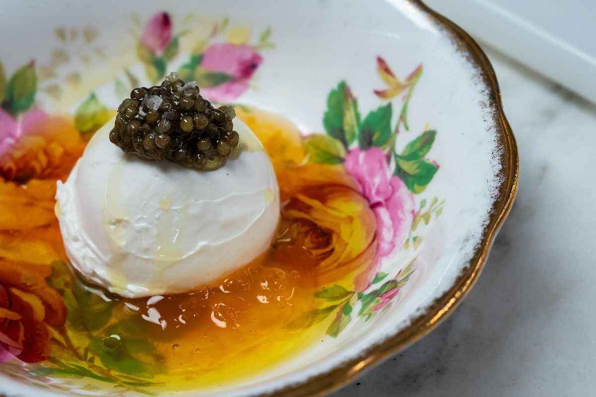 A white scoop of caviar-topped gelato sits on top of an orange sauce inside a floral bowl with gold rim.
