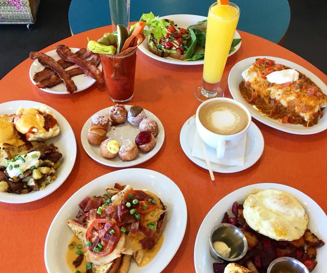 A close-up photo of a table covered with dishes containing assorted food from Jelly, including hashes, biscuits and gravy and salad, plus coffee, mimosas and bloody Marys in cups