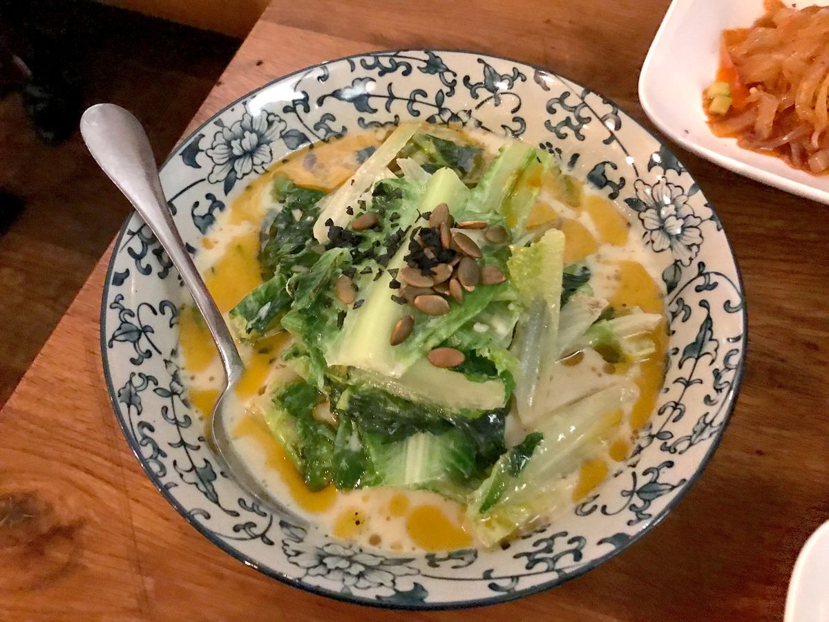 A bowl with romaine lettuce in soy milk broth topped with scallion oil and pumpkin seeds.