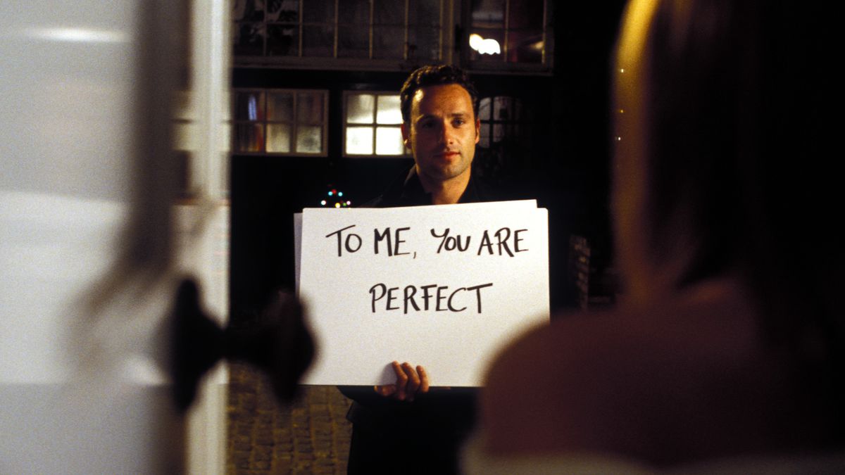 Mark (Andrew Lincoln) stands in a doorway, holding up a handwritten cue card that says “To me, you are perfect” in Love, Actually